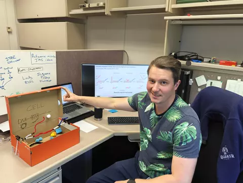 Sam Schaffter poses sitting in his office with a shoebox modified with craft supplies to be an educational model of an E. coli cell. 