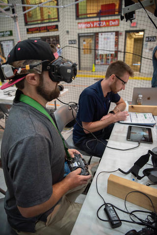 A man wearing an electronic device over his eyes sits at a table holding a controller for a drone. The indoor training facility includes pretend storefronts like a pizza shop. 