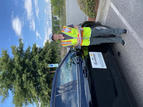 Jennifer Helgeson, holding a clipboard and wearing a high-vis vest stands next to a car. The car has a large sign on the side that reads “Center for Community Resilience Planning. Lumberton, NC Flood Study Team.” 