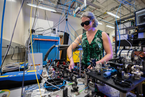 Alejandra Collopy dons safety goggles while she operates in the laboratory. She is encircled by various equipment and multiple hanging cables. 
