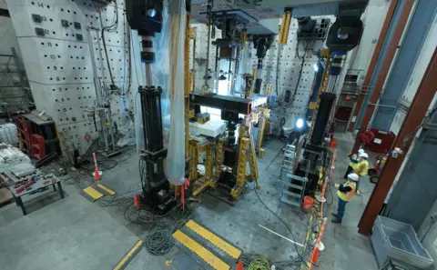 A high-ceilinged lab space is filled with large pieces of equipment, including a square structure in the center. A few people in hard hats stand at the edge of the room. 