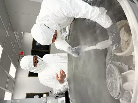 Researchers in white hazmat coveralls reach into an open freezer with a scooping device as vapor rises from the freezer.