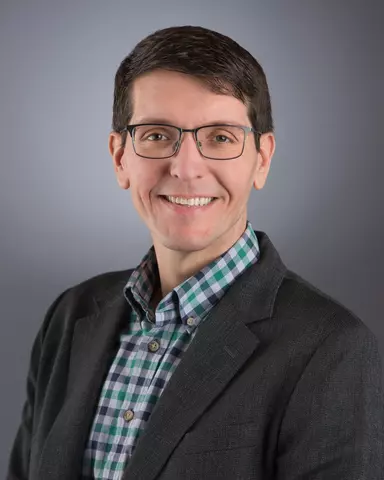 A headshot of Anthony Kotula wearing a checkered shirt, suit jacket, and glasses.