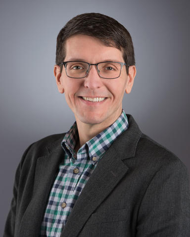 A headshot of Anthony Kotula wearing a checkered shirt, suit jacket, and glasses.