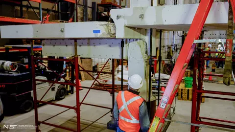 A person wearing construction safety gear looks up at a cracked concrete slab and beam arrangement held up by scaffolding in a large indoor space. 