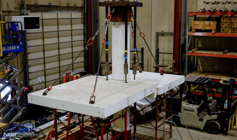 View from above shows cracked concrete slab attached by chains to column and held up from below by scaffolding in a large indoor space. 