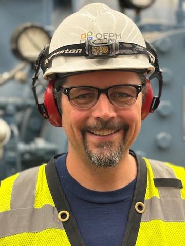 Man wearing a yellow safety vest and white construction helmet with ear protection and glasses smiles at the camera