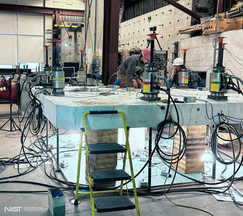 Two researchers in safety gear examine a slab of concrete set up horizontally in a large lab surrounded by scientific equipment.