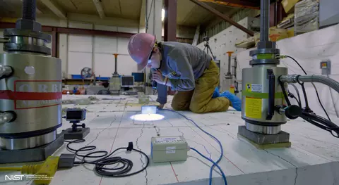 A researcher in a hard hat kneels on a concrete slab and uses a flashlight to examine it closely.