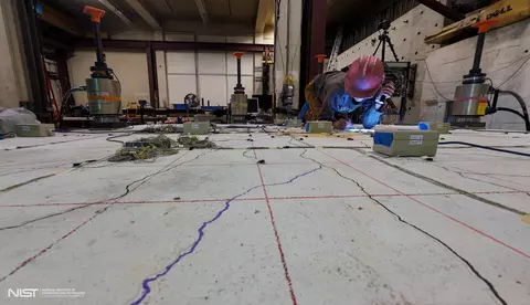 A researcher in a hard hat is making notes while kneeling on a concrete slab surrounded by scientific equipment.