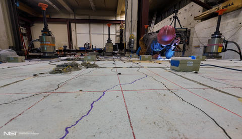 A researcher in a hard hat is making notes while kneeling on a concrete slab surrounded by scientific equipment.