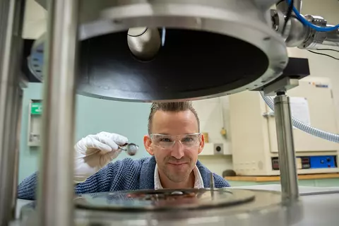 Andrew Iams wears safety glasses as his face is framed in an opening between two horizontal pieces of equipment in the lab. 