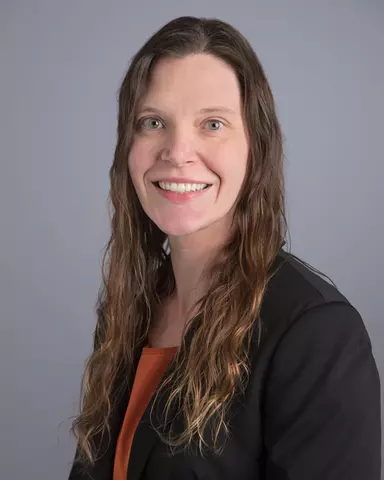 Professional portrait of Debra Audus, a woman with long wavy brown hair, smiling, wearing a black blazer.