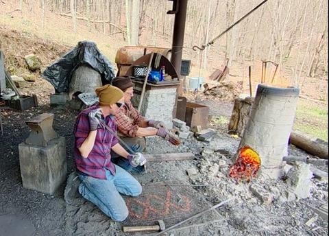 Andrew Iams kneels over a bloomery furnace in an outdoor, rural setting. 