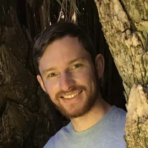 A photo of Tyler Laird in between two trees. His shoulders and head are visible and he is smiling and wearing a light blue shirt.