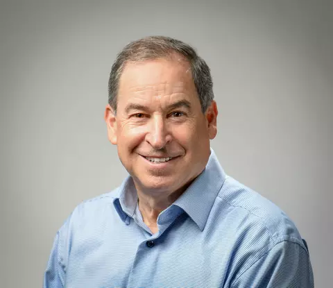 A man with short gray hair, wearing a blue collared shirt, smiling.