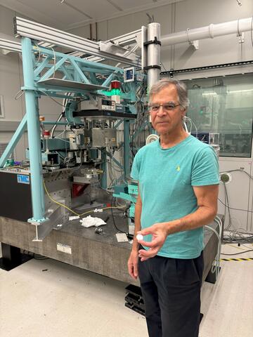 Jim Cline stands in front of a large scientific device in a lab, holding a small white disk.