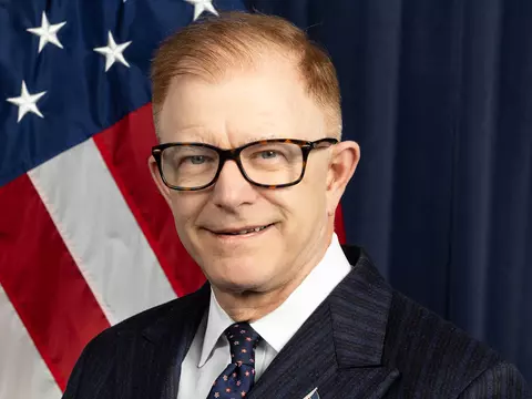 Headshot of Craig Burkhardt wearing glasses and a dark suit with an American flag behind him