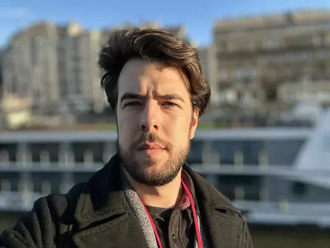 Headshot of August Staubus, a man with short brown hair, wearing a jacket, taking a selfie in front of a cityscape.