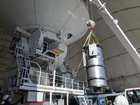 A large cylindrical device is being lifted by a crane next to the supporting structure of a telescope dish. 