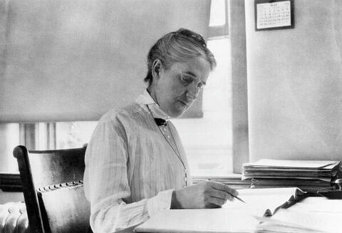 In a black-and-white photo, Henrietta Swan Leavitt sits at a desk, writing on a piece of paper.