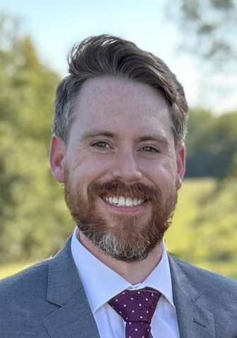 Headshot of Ian Hines, a man with short hair, mustache, and beard, wearing a suit and tie, smiling for a photo outdoors.