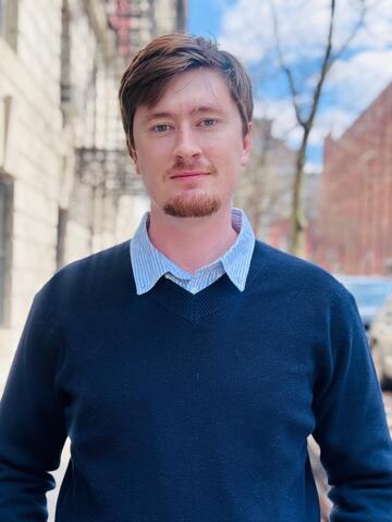 A photo of Benjamin Eller, wearing a blue sweater and smiling casually, framed from the midsection up to the head. 