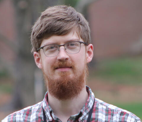 Headshot of Michael Haines, a man with short hair, mustache, beard, and glasses, wearing a collared plaid shirt, posing for a photo in front of a tree.