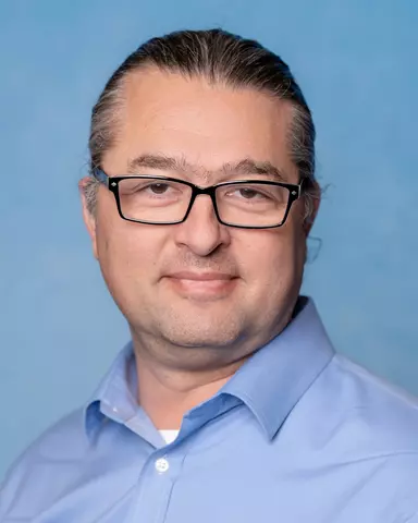 Jason Kralj, a man with long dark hair pulled back into a ponytail and wearing glasses, smiling for a professional headshot. 