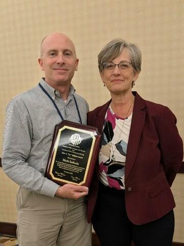 NIST's Mark Iadicola, on the left, and ASTM E28 Chair Sue Marvin, on the right, posing together, with the Millane Award, a wooden plaque. The plaque features a black surface with gold lettering, displaying the ASTM logo, Mark Iadicola's name, and other inscribed details. 