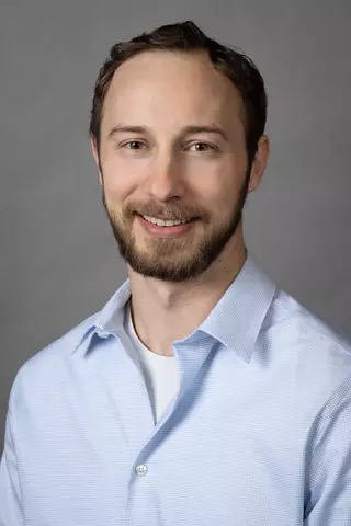 Professional portrait of Newell Moser, a man with short dark hair and beard, smiling, wearing a blue collared shirt.