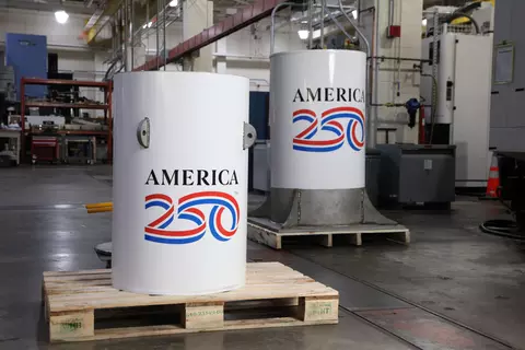 Two white cylinders with the America 250 logo stand in a large workshop space. 