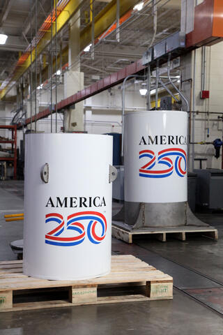 Two white cylinders with the America 250 logo stand in a large workshop space.
