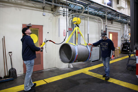 Two men use an overhead pulley apparatus to move a thick metal cylinder inside a large workshop. 