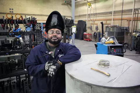 A man wearing an uplifted welding mask and safety glasses poses leaning on a large metal cylinder in a workshop. 