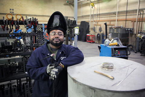 A man wearing an uplifted welding mask and safety glasses poses leaning on a large metal cylinder in a workshop. 