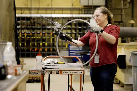 A woman wearing safety glasses holds an electric tool to work on a circle of metal in a large workshop. 