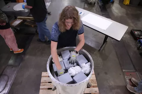 An overhead view shows a woman reaching into a cylindrical container that holds several gray boxes along with what looks like crumpled paper. 