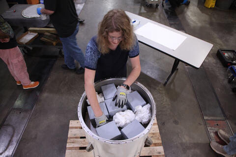 An overhead view shows a woman reaching into a cylindrical container that holds several gray boxes along with what looks like crumpled paper. 