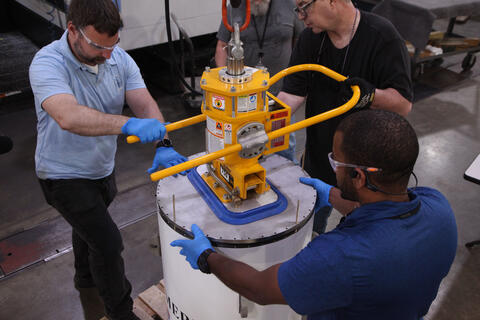 Three men stand around the cylindrical time capsule, handling a yellow device that holds the lid of the cylinder.