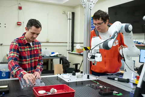Two young men stand a lab table fitted with a white articulated robot arm.