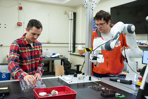 Two young men stand a lab table fitted with a white articulated robot arm.