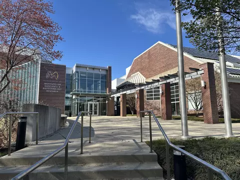 Concrete stairs with metal railings lead up to a sunny courtyard outside a modern campus building. The entrance to this building is decorated with tall glass windows and dark bricks. To its left is a logo for the Institute for Bioscience and Biotechnology Research (IBBR). To the right, there is a larger brick building with a sloped roof that has solar panels, and a wooden walkway structure. The scene is brightly lit by the clear blue sky.