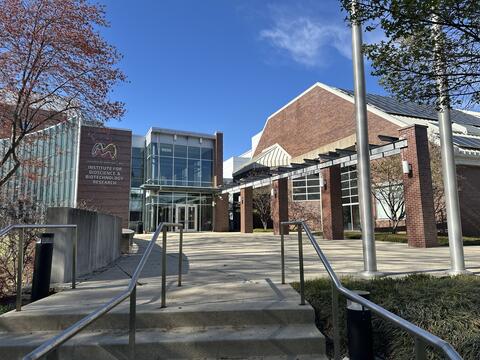 Concrete stairs with metal railings lead up to a sunny courtyard outside a modern campus building. The entrance to this building is decorated with tall glass windows and dark bricks. To its left is a logo for the Institute for Bioscience and Biotechnology Research (IBBR). To the right, there is a larger brick building with a sloped roof that has solar panels, and a wooden walkway structure. The scene is brightly lit by the clear blue sky.
