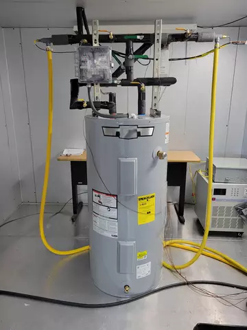 A water heater sits in a lab with several pipes and wires coming out of the top. 