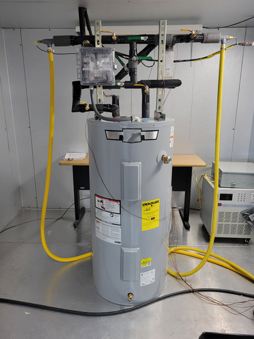 A water heater sits in a lab with several pipes and wires coming out of the top. 