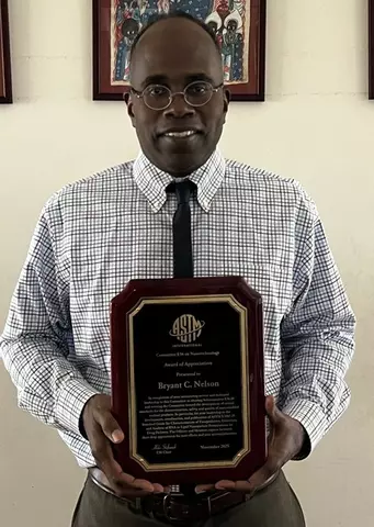 Bryant C. Nelson, a man wearing glasses, a checkered dress shirt, and a tie, smiling while holding his ASTM International Committee E56 on Nanotechnology Award of Appreciation Plaque. The wooden plaque features the ASTM logo and details about the award including the recipient's name, the E56 Chair's signature, and "November 2025" engraved in gold against a black background. 