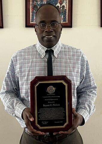Bryant C. Nelson, a man wearing glasses, a checkered dress shirt, and a tie, smiling while holding his ASTM International Committee E56 on Nanotechnology Award of Appreciation Plaque. The wooden plaque features the ASTM logo and details about the award including the recipient's name, the E56 Chair's signature, and "November 2025" engraved in gold against a black background. 