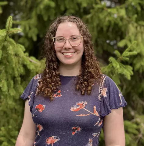 Kristen Ramsey (a woman in a blue shirt) stands in front of green pine trees in a professional headshot