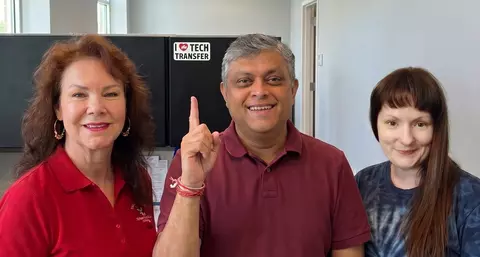 From left to right, Cecelia Foote, Saikat Kumar Ghosh, and J'aime Maynard smiling and posing for a group photo in an office, with Saikat Kumar Ghosh pointing to a "I (Heart) Tech Transfer" Sign in the background behind them. 